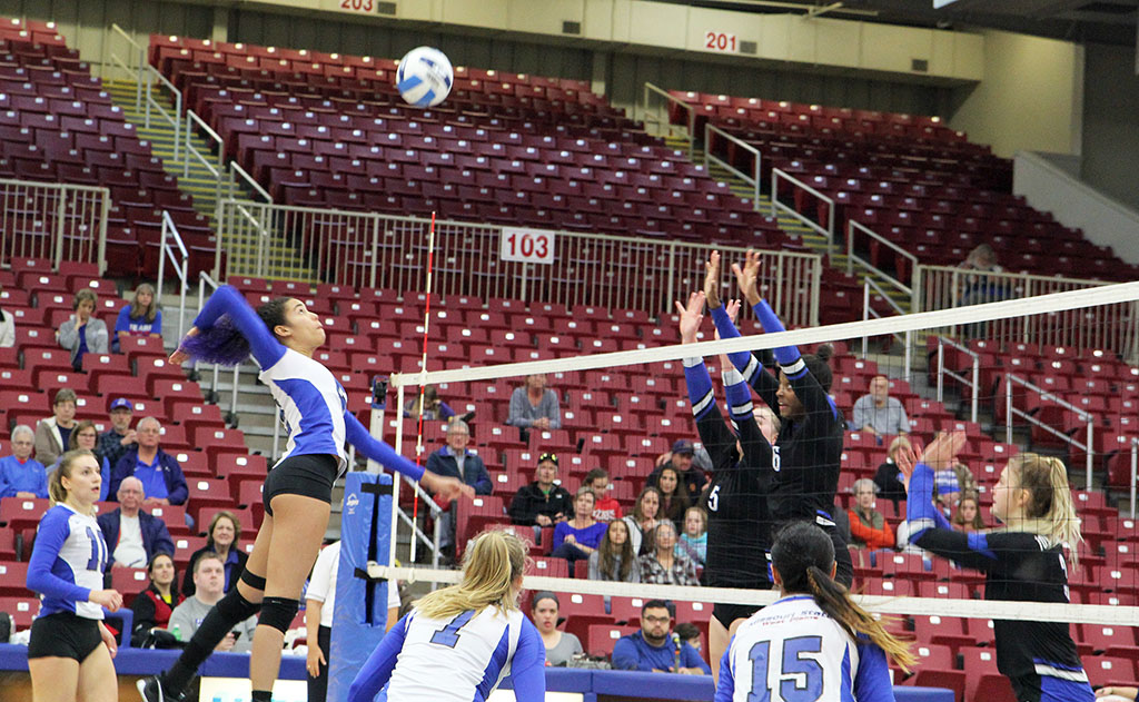 A volleyball player jumps into the air to hit the ball while her teammates look on, and her opponents jump up across the net to try to block the ball.