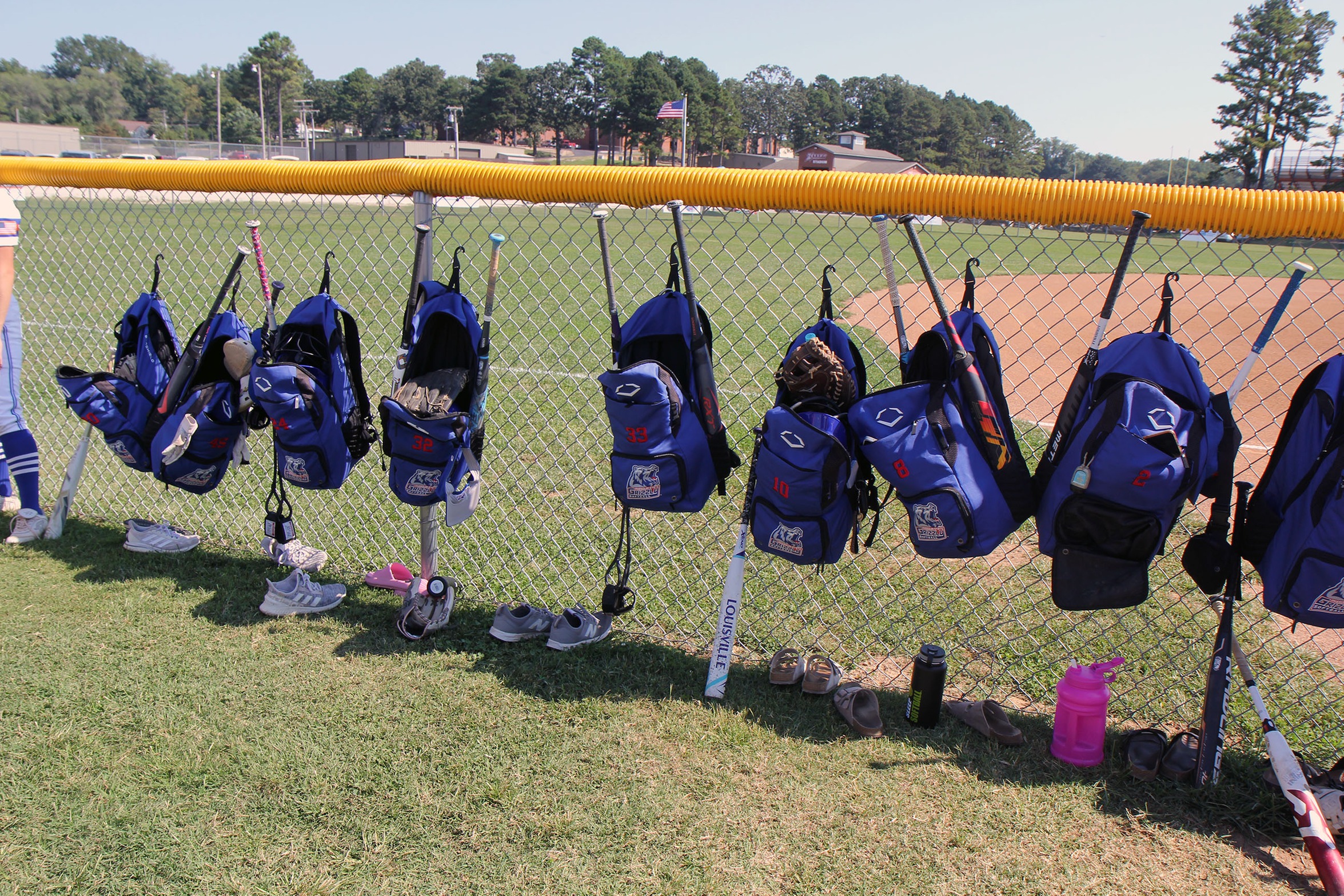 Backpacks filled with softball gear hang on a chainlink fence.